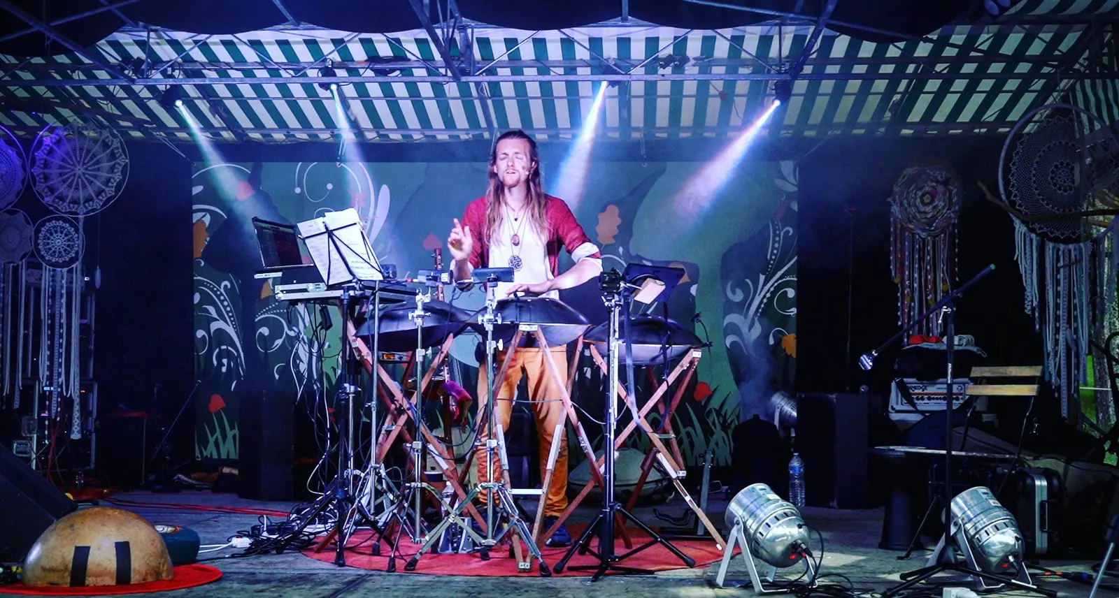 David Lesage standing on stage under the spotlights, behind his two handpans mounted on stands, surrounded by his stands, his pedals and a calabash resting on the red carpet.