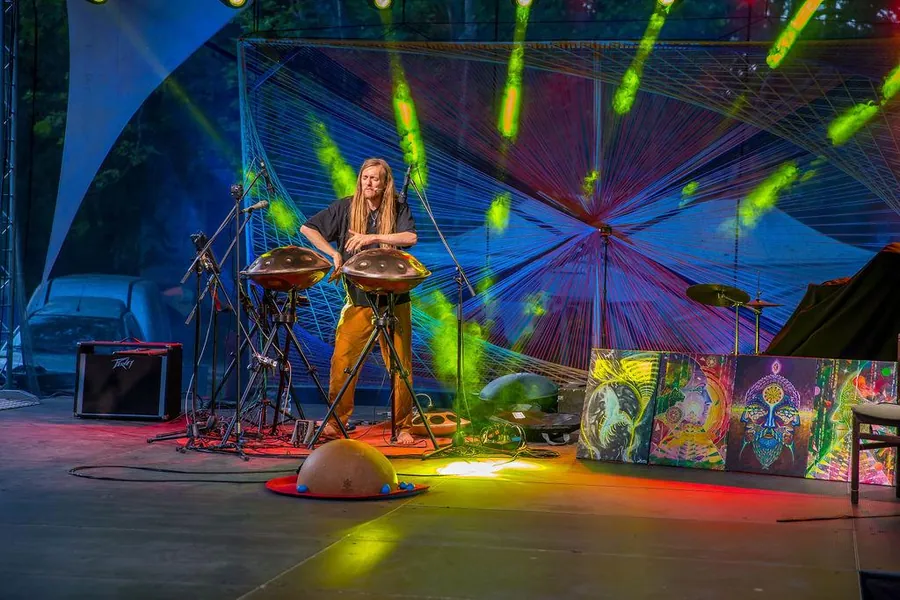 David Lesage alone on the stage of the Everness Festival in Hungary: standing barefoot behind his two acoustic handpans mounted on black tripods, under green and red light beams, in front of a large multicoloured woven backdrop; a calabash half-sphere rests on the floor in front of him.