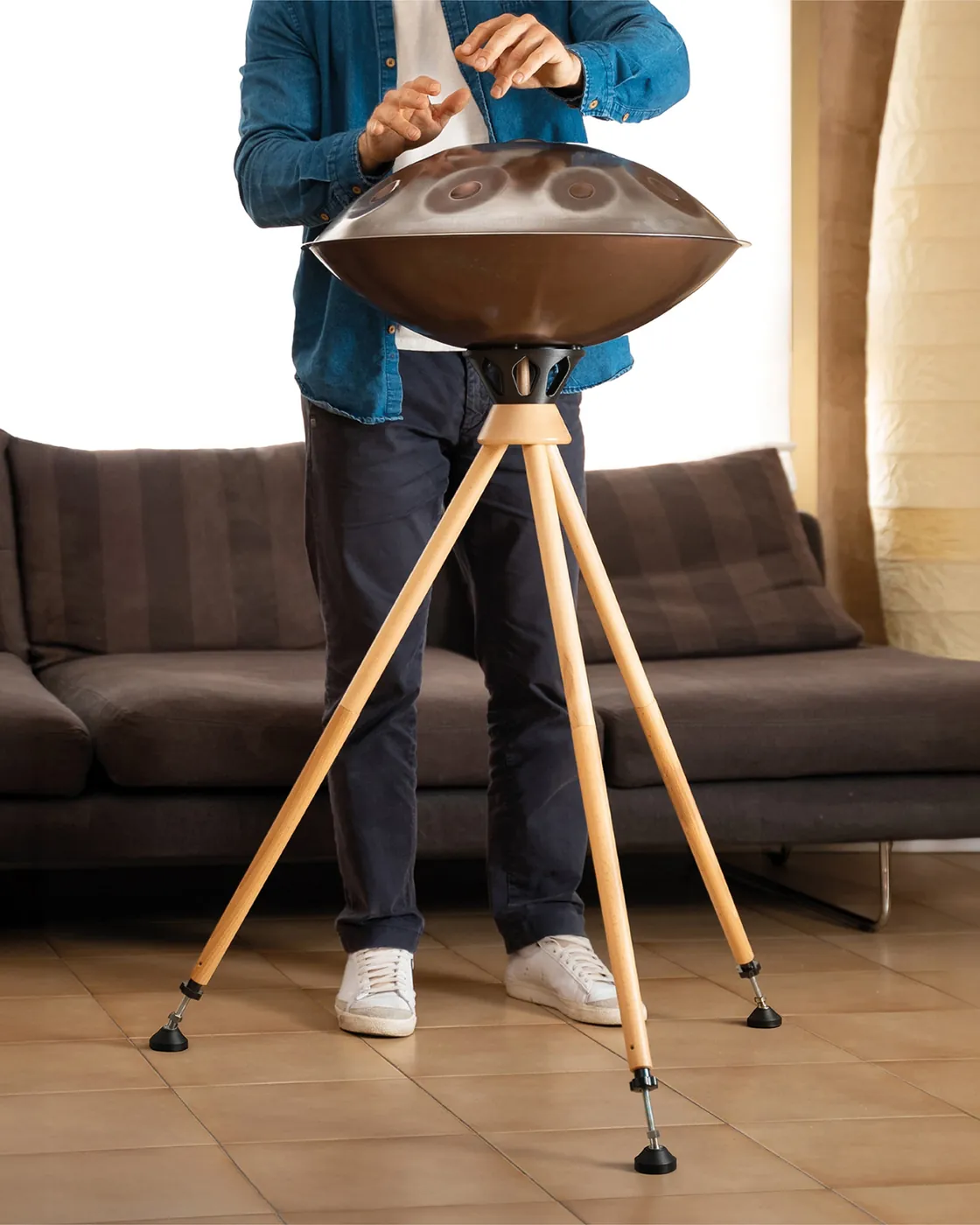 A musician plays standing at a handpan resting on the Atlas All tripod, in a living room: the three light wooden legs are extended to the high position.