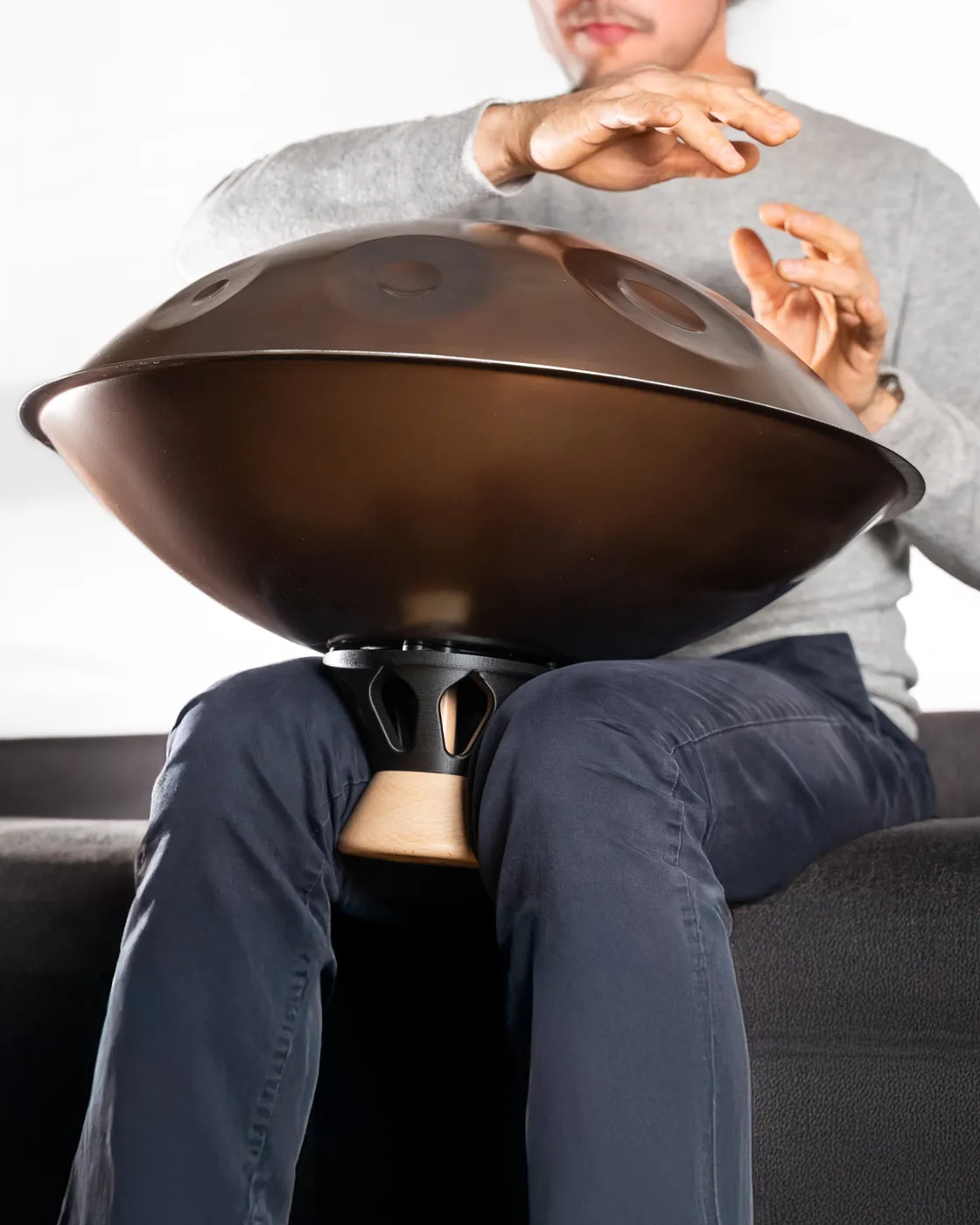 A person seated on a sofa plays a handpan held by the Atlas Body gripped between their thighs: the instrument is lifted clear of their legs.