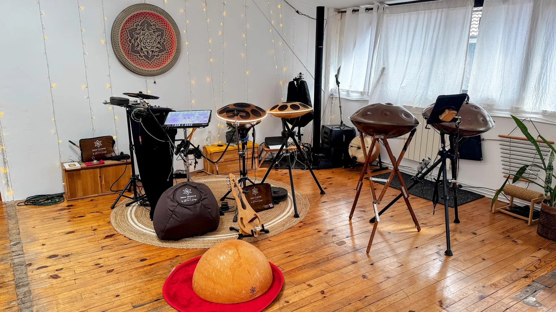 David Lesage’s showroom set up before a showcase: two Neotone electronic handpans with a lit rim, on black tripods next to an electronic drum kit, and two Yishama acoustic steel handpans, one on a wooden tripod, the other on a metal stand; a calabash half-sphere resting on a red mat in the foreground, old wooden floor, fairy lights and a rosette on the wall.